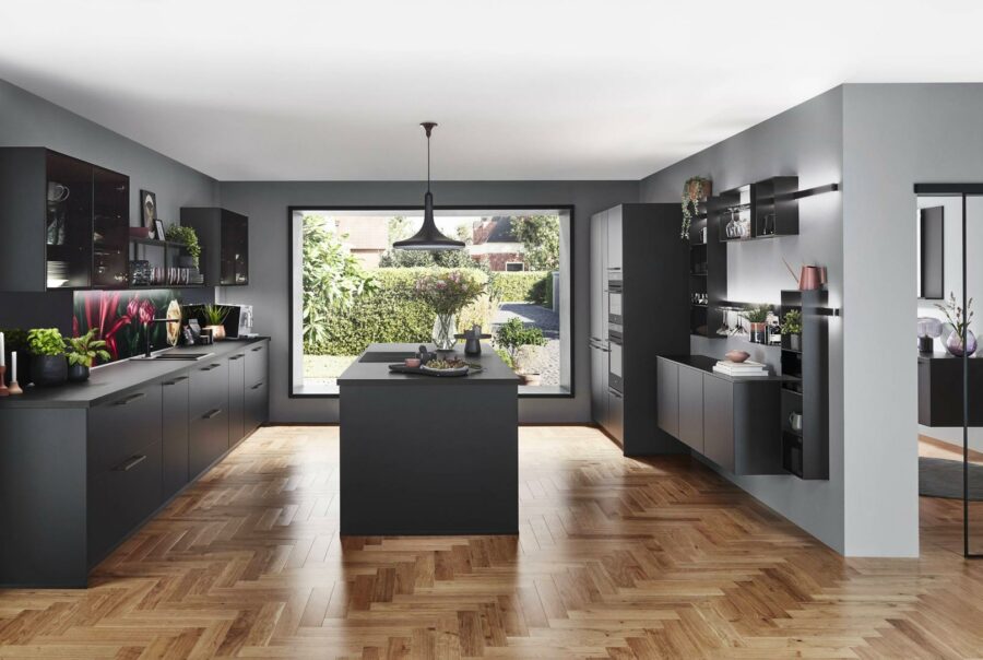 Cuisine moderne avec îlot central noir, parquet en bois, murs gris et grande baie vitrée donnant sur jardin verdoyant
