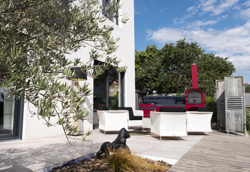 Terrasse moderne avec salon de jardin blanc, four à pizza rouge et olivier, chien noir se reposant au soleil