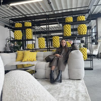 Femme assise sur canapé beige dans showroom moderne avec étagères métalliques remplies de boules jaunes décoratives
