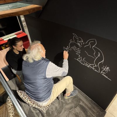 Homme âgé dessinant un personnage de dessin animé sur un mur noir tandis qu'une femme le regarde depuis un escalier