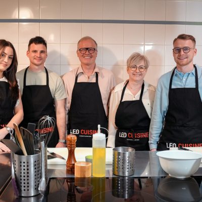 Cinq personnes en tabliers noirs Leo Cuisine posent souriantes dans une cuisine professionnelle moderne
