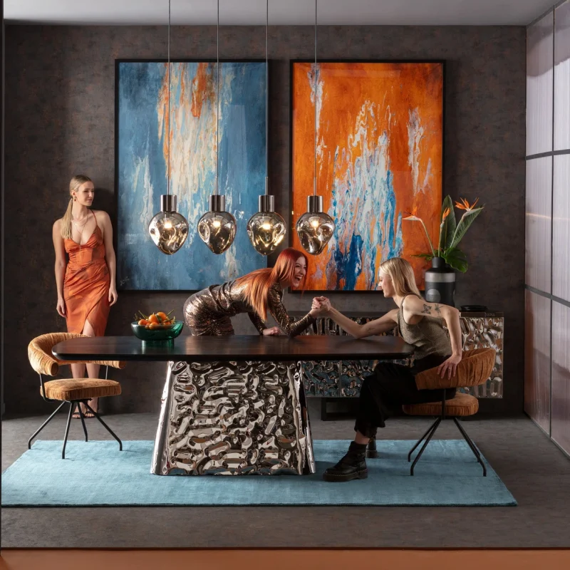 Trois femmes dans une salle à manger moderne avec table sculptée, suspensions dorées et tableaux abstraits colorés