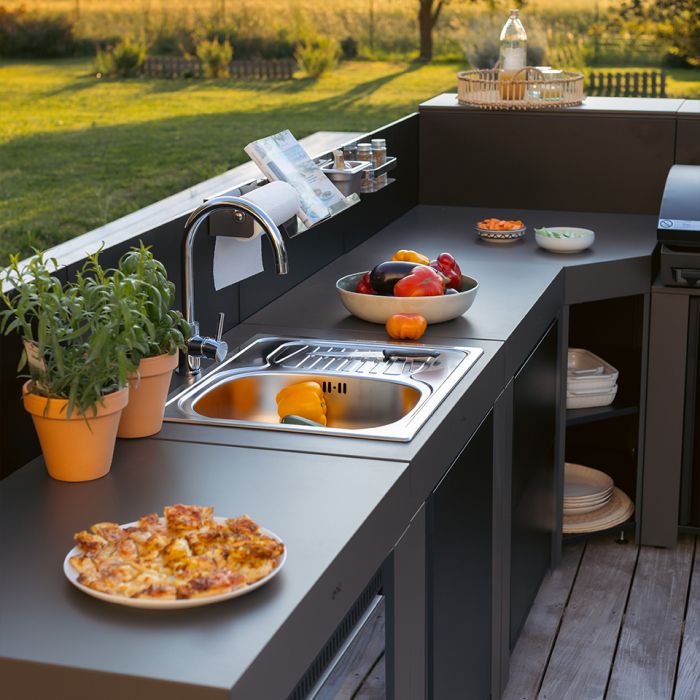Cuisine extérieure moderne avec évier, plan de travail noir, pizza, fruits et plantes aromatiques dans un jardin