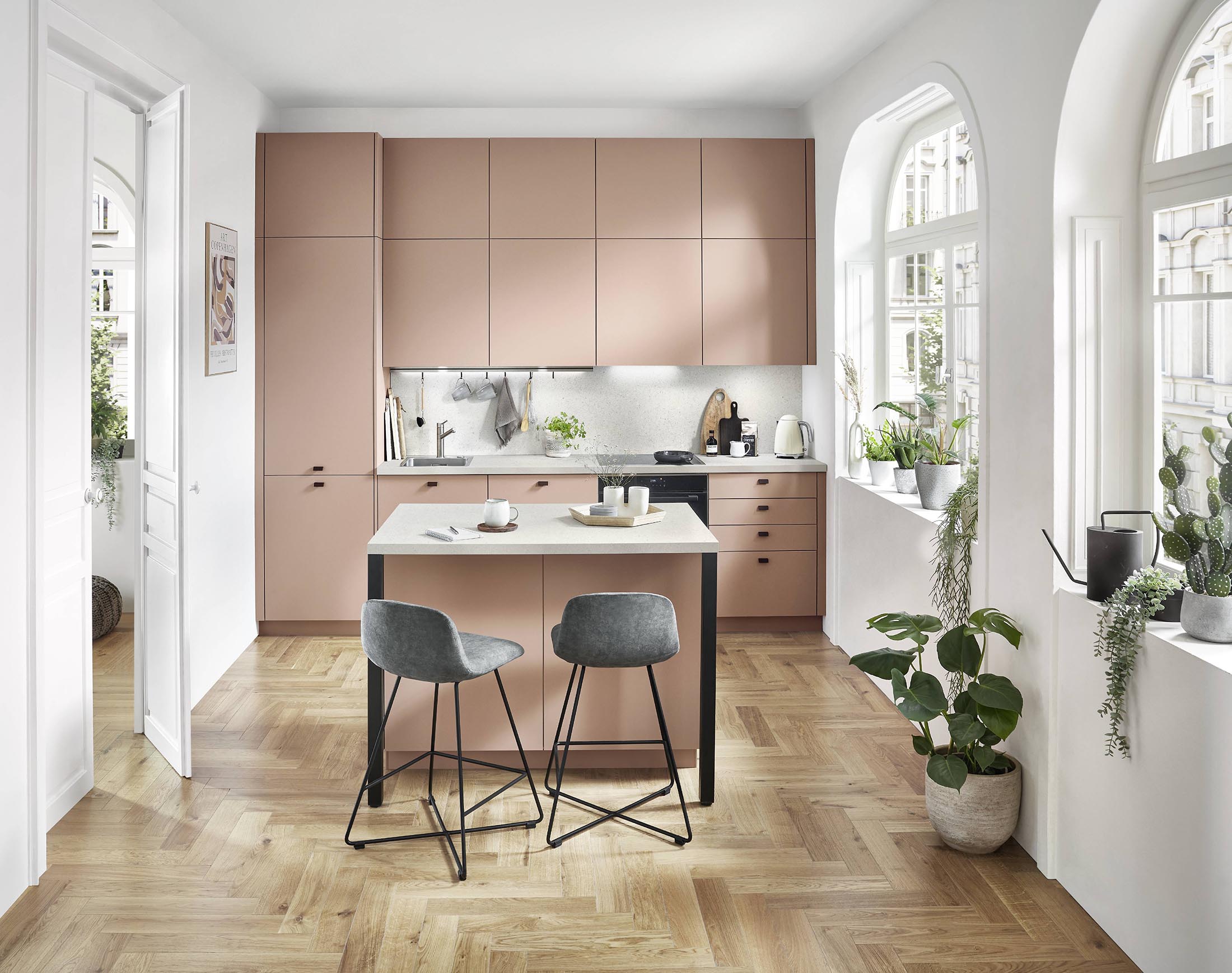 Cuisine moderne avec façades roses, îlot central, tabourets gris et grandes fenêtres dans appartement lumineux