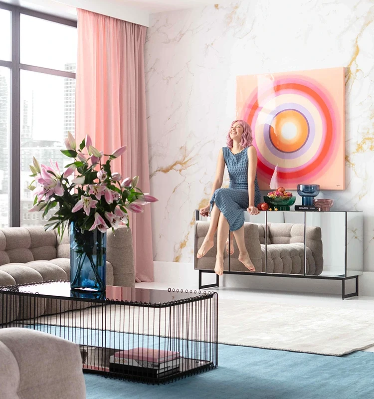Femme en robe bleue assise sur meuble miroir dans salon moderne avec artwork coloré, tapis bleu et rideaux roses